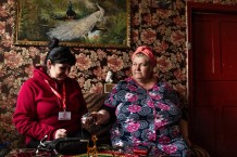Lubov, 57, a beneficiary, during a home visit with local community worker Olena, 55. Lubov has two biological children and nine adopted children, all orphaned and disabled. Olena is a widow and has a 24-year-old son who has been in the war since he was 19 and is now, after four years, the commander of his unit. Prygozhe, population 160, Kharkiv Region. Ukraine, 2026
