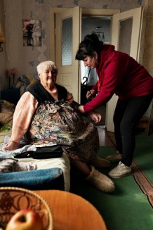 Valentina, 86, a beneficiary, during a home visit with local community worker Olena, 55. Valentina hasn't walked for a year. She lives with her daughter Antonina, 66, also a beneficiary. Her husband, son, and son-in-law have died, and her grandchildren have left the country. Prygozhe, population 160, Kharkiv Region. Ukraine, 2026